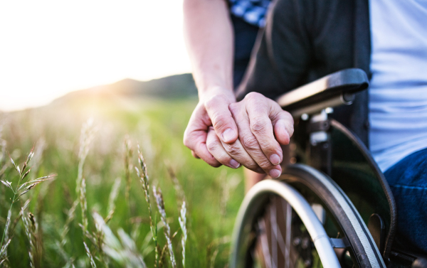 A close-up of unrecognizable son holding his father's hand on a wheelchair. Copy space.