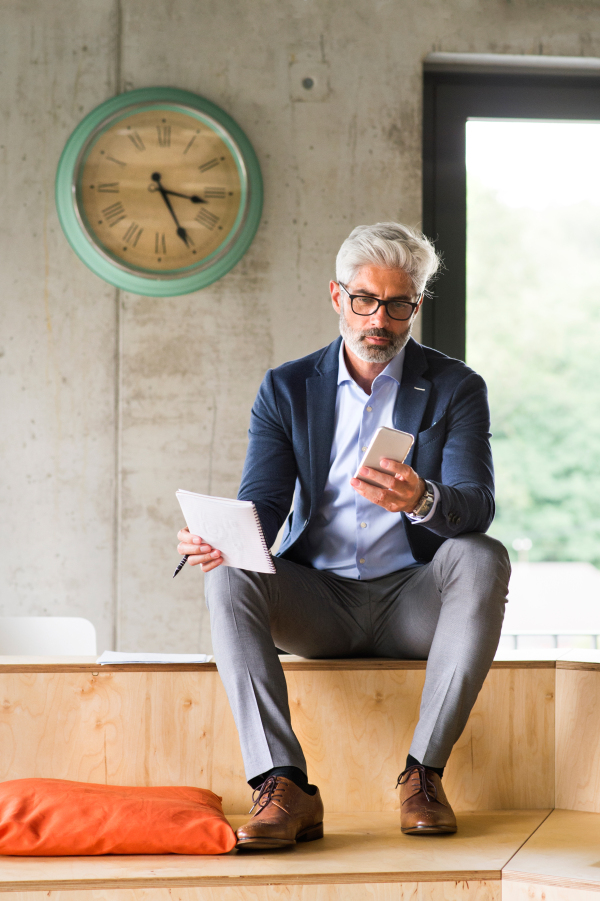 Thoughtful mature businessman with smartphone in the office, sitting on desk.