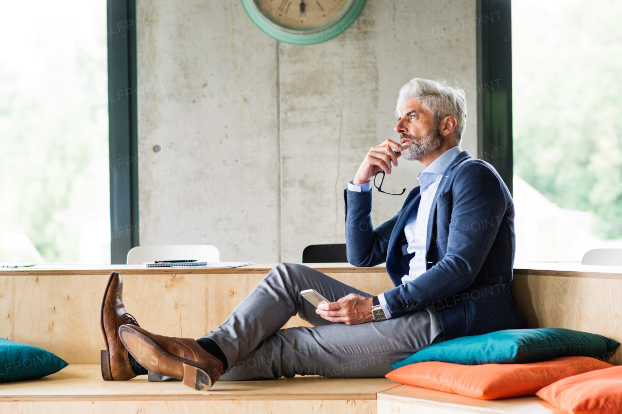 Thoughtful mature businessman with smartphone in the office, sitting with legs on desk.