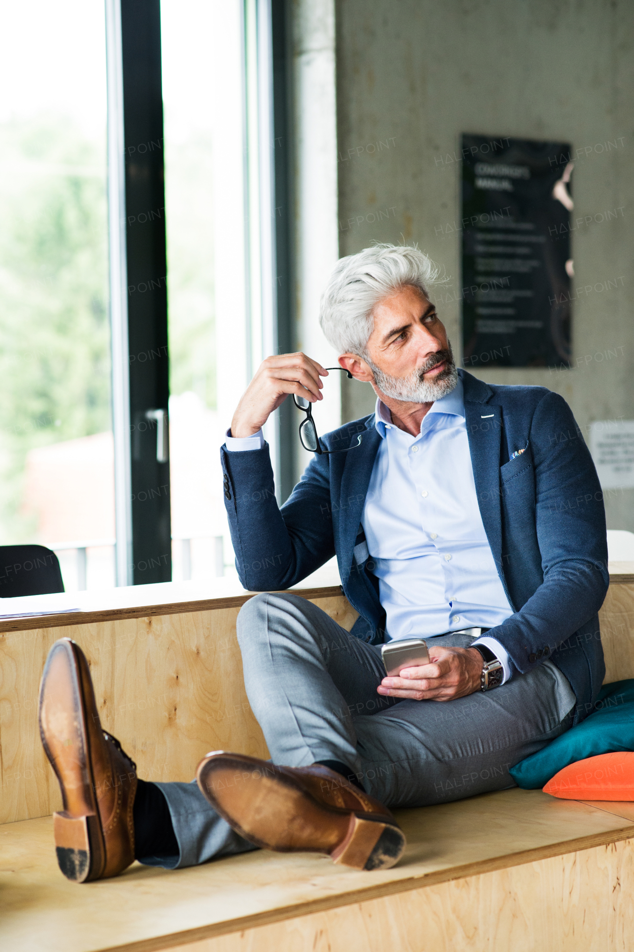 Thoughtful mature businessman with gray hair in the office wearing a blue shirt sitting with legs on desk.