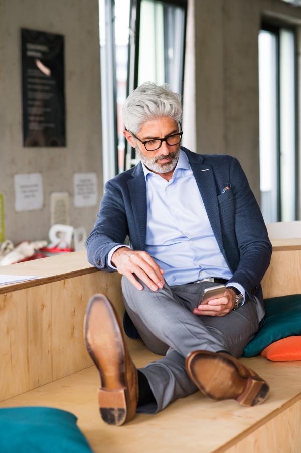 Thoughtful mature businessman with smartphone in the office, sitting with legs on desk.