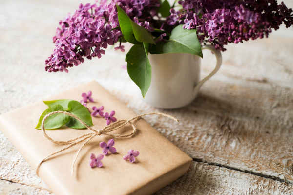 Beautiful purple lilac bouquet in vase laid on table and present wrapped in brown paper. Studio shot on white wooden background.