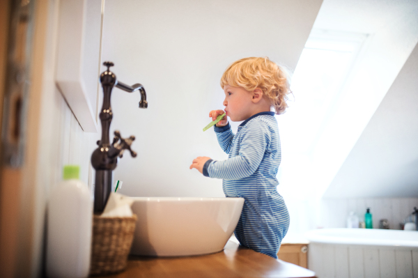 Cute toddler brushing his teeth in the bathroom. Little boy standing on a stool in front of mirror.