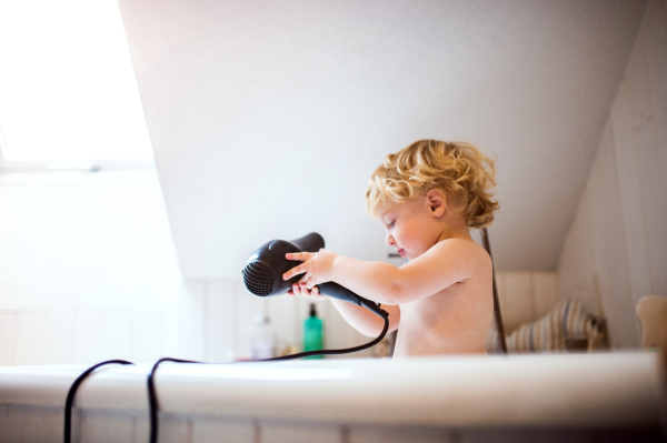 Cute little toddler boy with a hairdryer standing in the tub in the bathroom. Dangerous situation. Domestic accident.