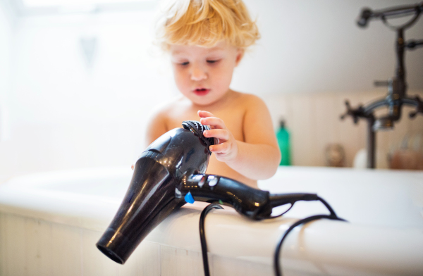 Cute little toddler boy with a hairdryer standing in the tub in the bathroom. Dangerous situation. Domestic accident.