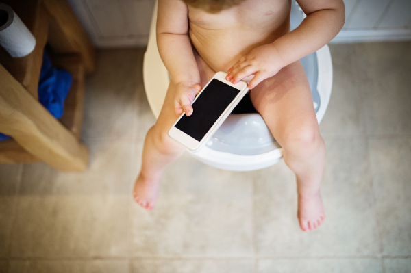 Unrecognizable toddler with a smartphone in the bathroom. Little boy sitting on the toilet. Top view.