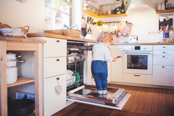 Little toddler boy standing on the dishwasher door. Domestic accident. Dangerous situation at home. Child safety concept.