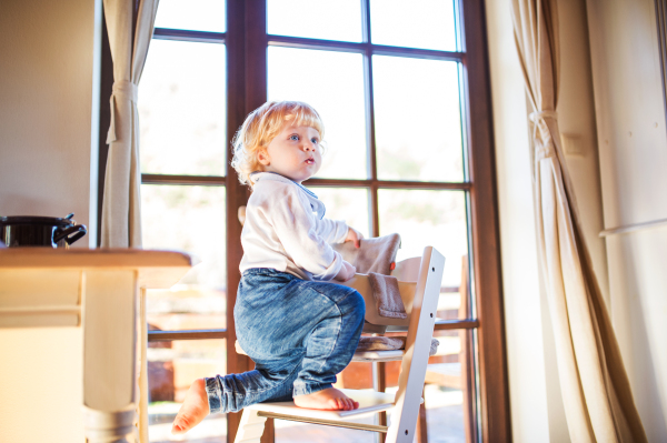 Little toddler boy climbing on wooden highchair at home. Domestic accident. Dangerous situation at home.