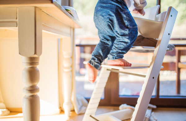 Little toddler boy climbing on wooden highchair at home. Domestic accident. Dangerous situation at home.