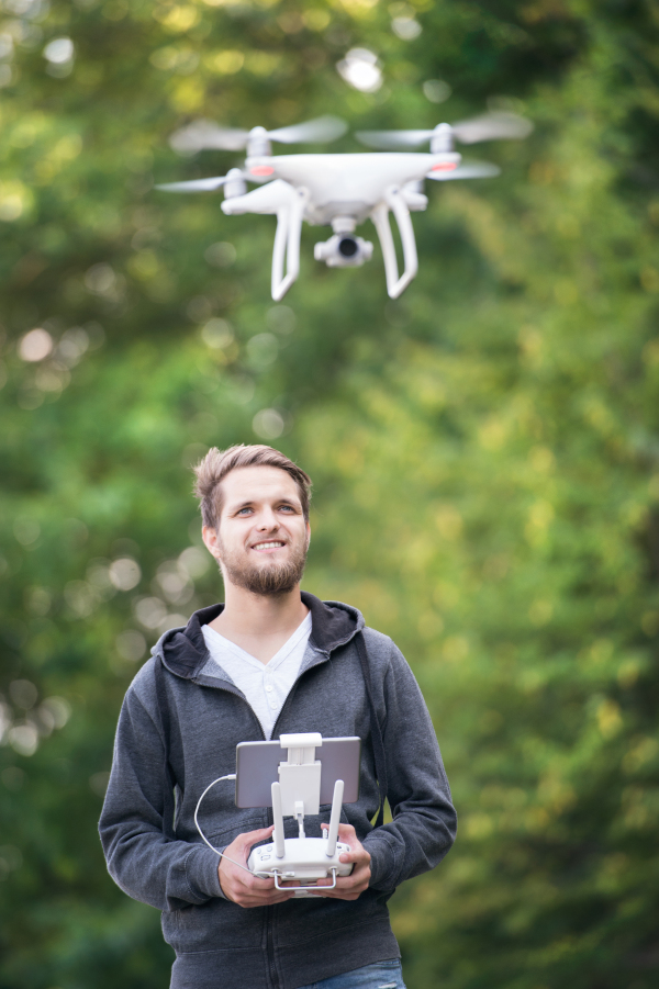 Young hipster man with remote control and flying drone with camera. Green sunny nature.