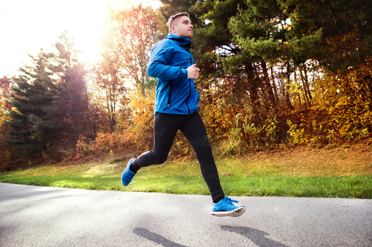 Young athlete in blue jacket running outside in colorful sunny autumn nature. Trail runner training for cross country running.