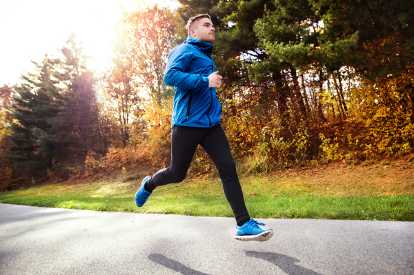 Young athlete in blue jacket running outside in colorful sunny autumn nature. Trail runner training for cross country running.