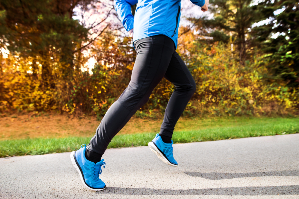 Unrecognizable young athlete in blue jacket running outside in colorful sunny autumn nature. Trail runner training for cross country running.