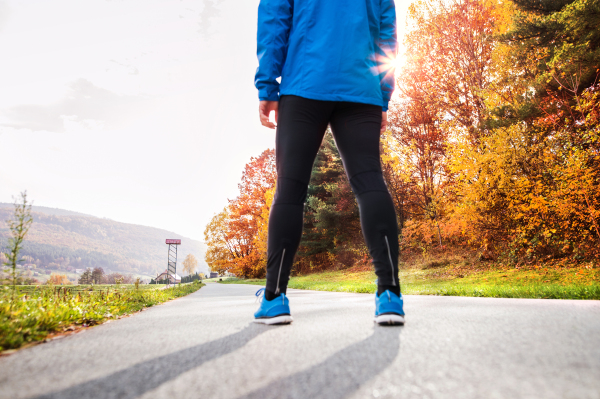 Unrecognizable young runner in blue jacket outside in colorful sunny autumn nature standing on an asphalt path. Trail runner training for cross country running. Rear view.