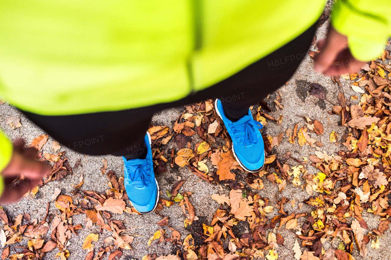 Unrecognizable young runner in yellow jacket outside in colorful sunny autumn nature standing on an asphalt path. Trail runner training for cross country running.