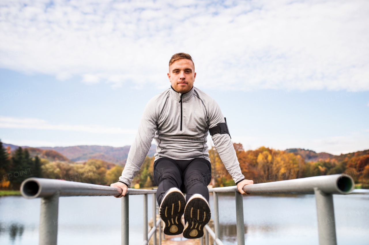 Young man with smartphone in gray jacket exercising outside in autumn nature. Parallel bars L-sit.