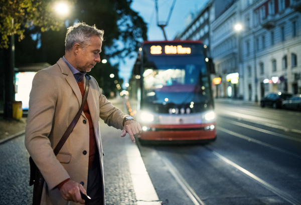 Mature businessman with suitcase waiting for a tram or a trolley car in the evening in Prague city, checking the time.