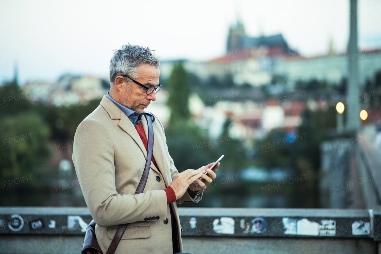 Mature businessman with smartphone standing on a bridge in city, text messaging.