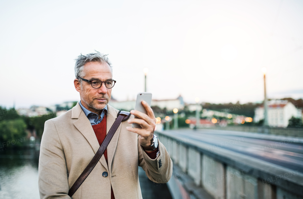 Mature handsome businessman with smartphone standing by river Vltava in Prague city, taking selfie. Copy space.