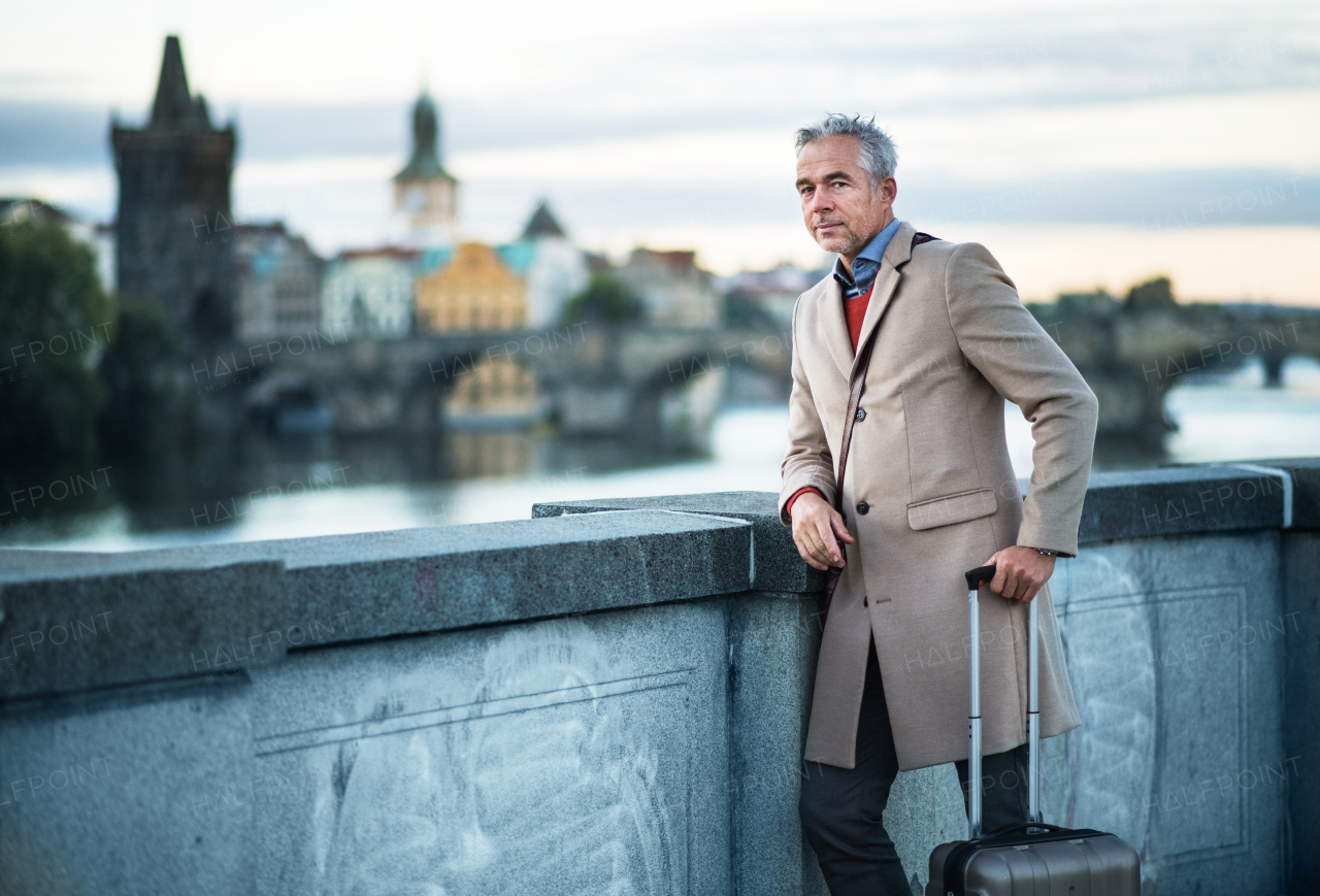 Mature handsome businessman with suitcase standing on a bridge in Prague city. Copy space.