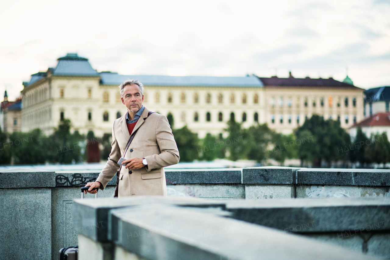 Mature handsome businessman with suitcase and smartphone standing on a bridge in Prague city. Copy space.