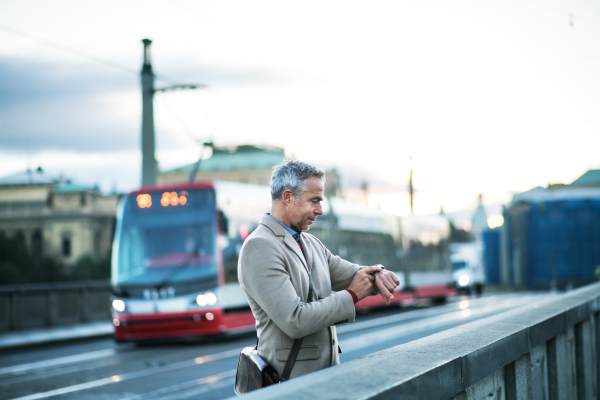 Mature handsome businessman with smartwatch standing on a bridge in Prague city, checking the time. Copy space.