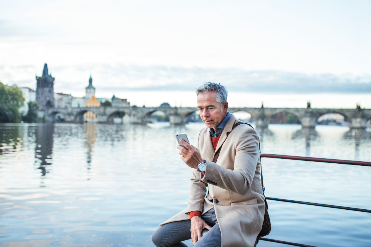 Mature handsome businessman with smartphone sitting by river Vltava in Prague city. Copy space.