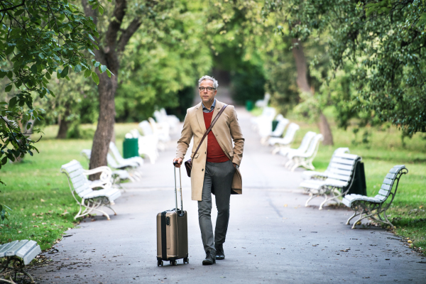 Mature handsome businessman with suitcase walking in a park in a city.