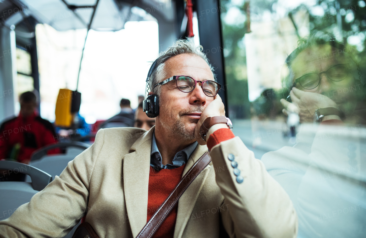 Mature tired businessman with glasses and heaphones travellling by bus in city, listening to music.