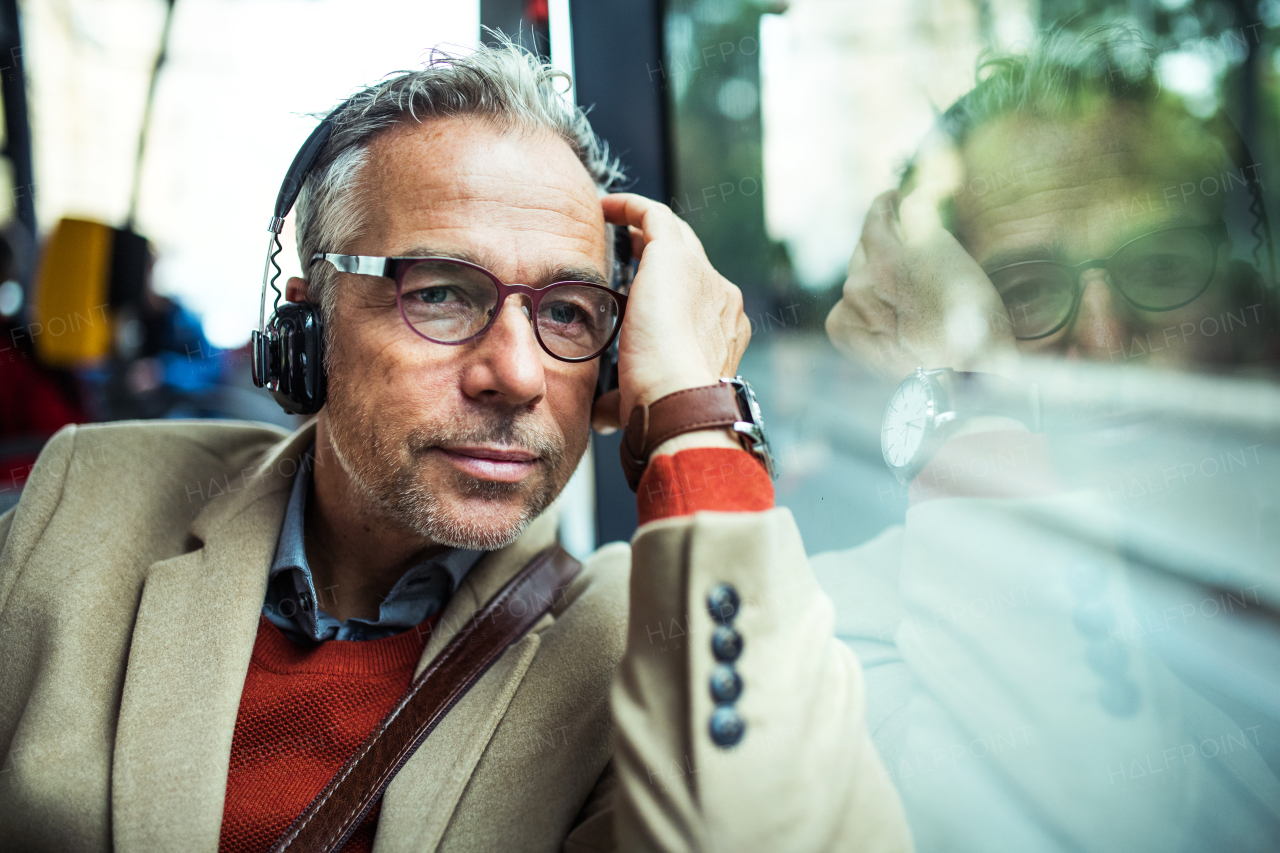 Mature tired businessman with glasses and heaphones travellling by bus in city, listening to music.