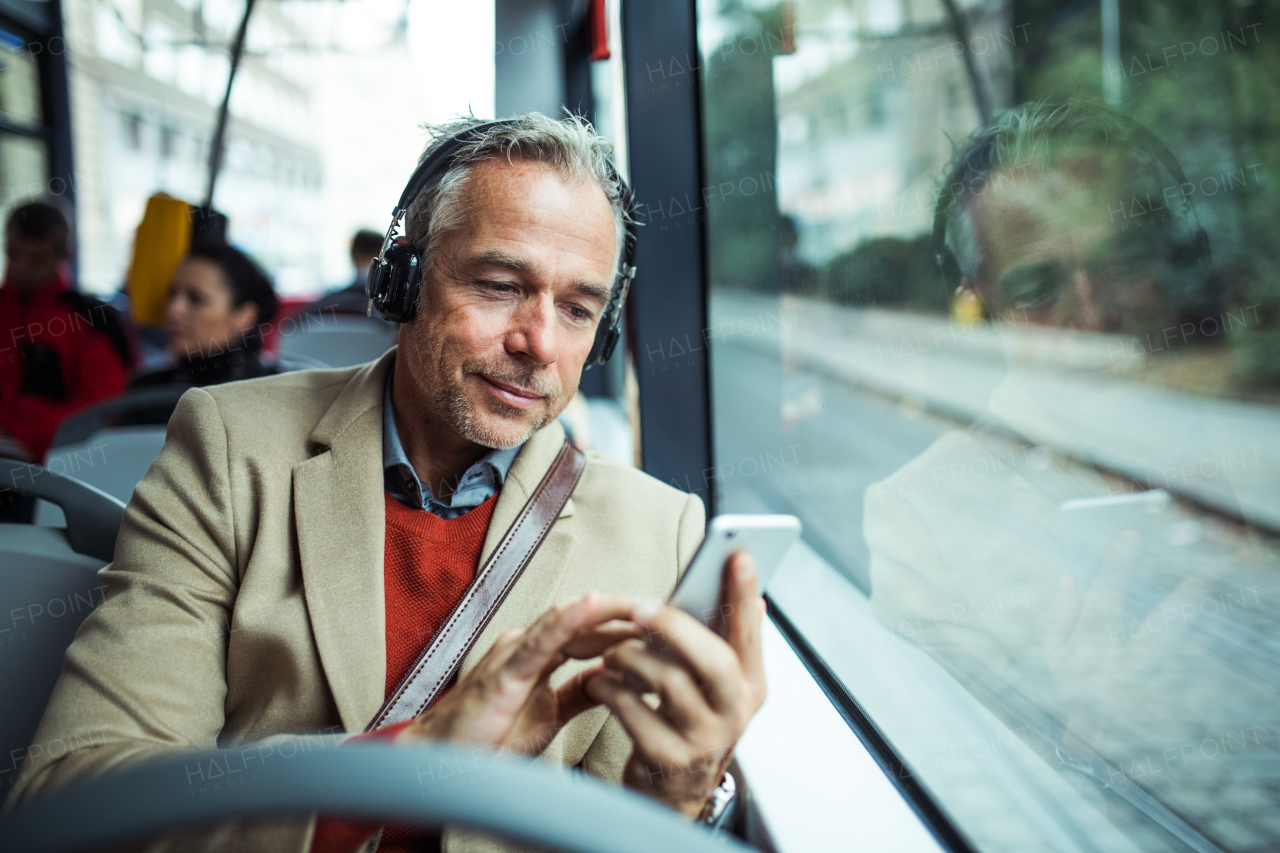 Mature tired businessman with smartphone and heaphones travellling by bus in city, listening to music.