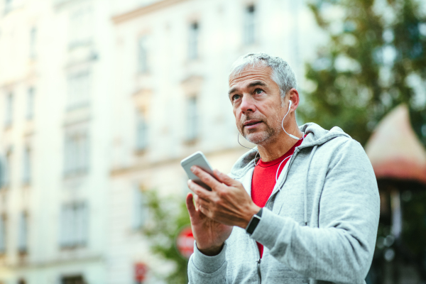 A portrait of an active mature man with earphones standing outdoors in city, using smartphone.