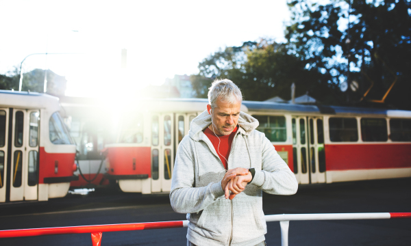 A portrait of an active mature man with earphones standing outdoors in city, checking the time.