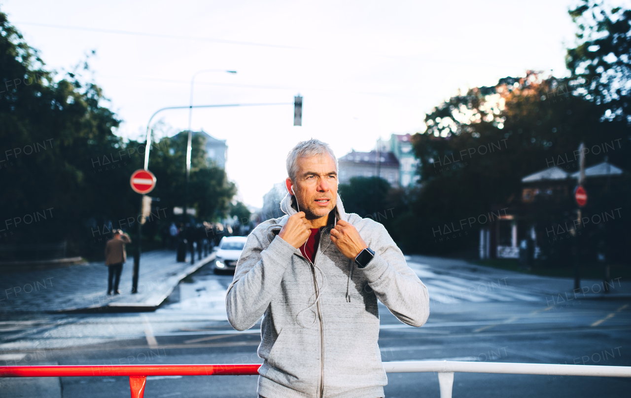 A portrait of an active mature man standing outdoors in city, putting earphones in his ears.