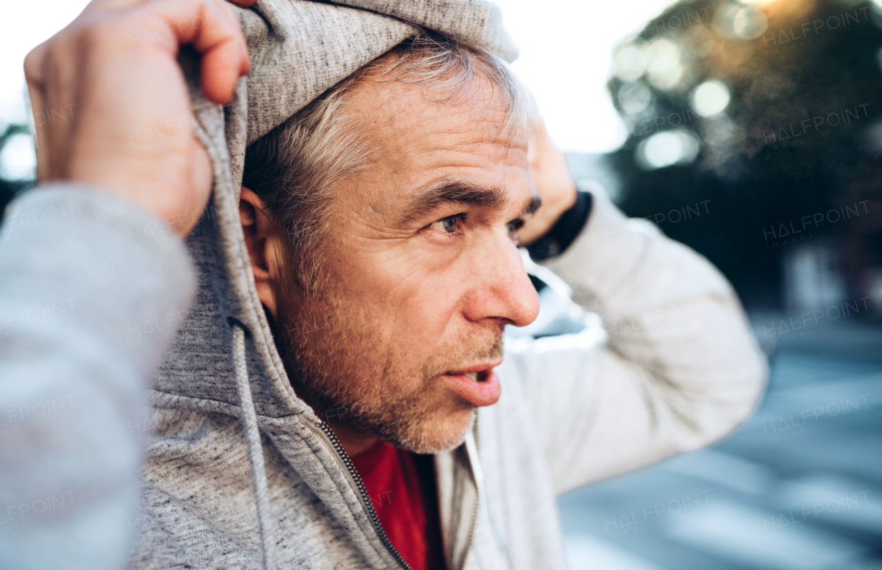 A portrait of an active happy mature man standing outdoors in city, putting gray hood on head.