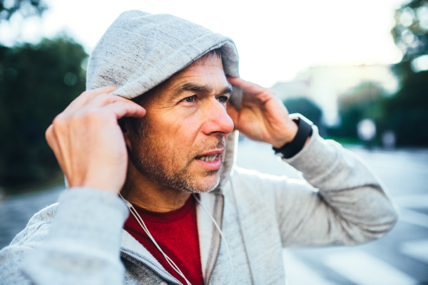 A portrait of an active happy mature man with earphones standing outdoors in city, putting gray hood on head.