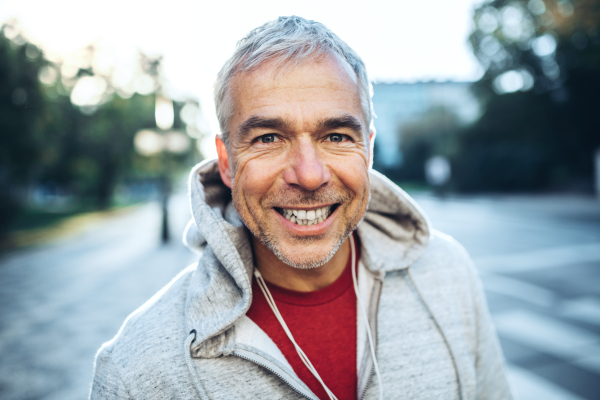 A portrait of an active happy mature man standing outdoors in city, wearing gray hoodie.