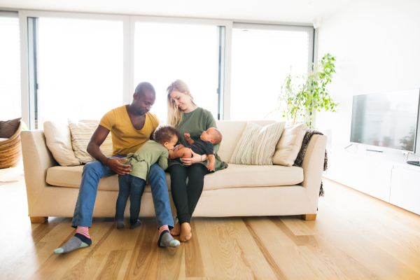 Beautiful young interracial family at home with their cute daughter and little baby son, sitting on couch.