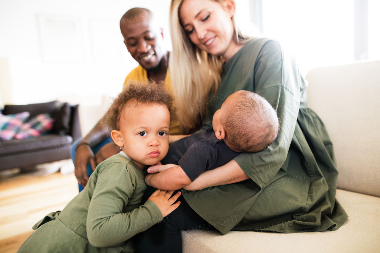 Beautiful young interracial family at home with their cute daughter and little baby son, sitting on couch.