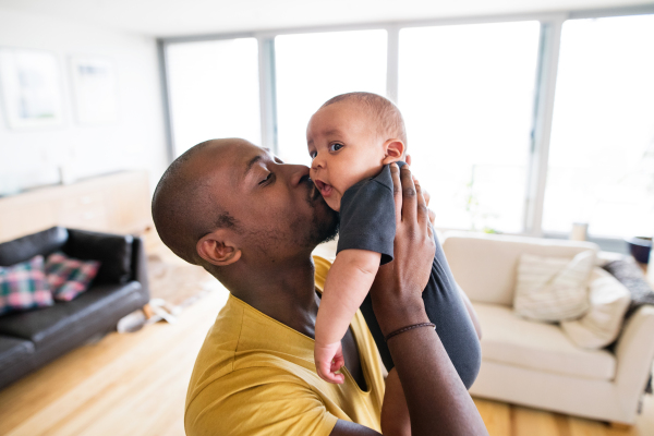 Young afro-american father at home holding his cute baby son in his arms, kissing him.