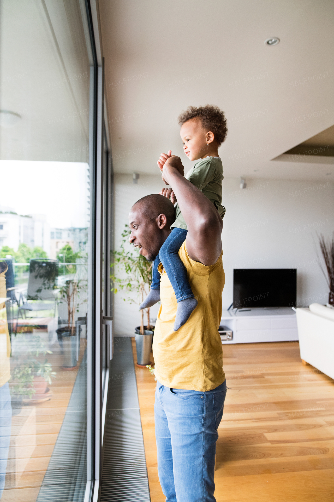 Young afro-american father at home at the window carrying his little daughter on his shoulders.