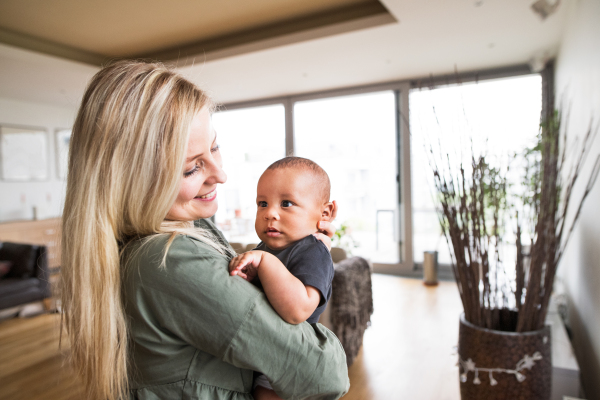 Beautiful young blonde mother at home holding her little mixed-race baby son in the arms.