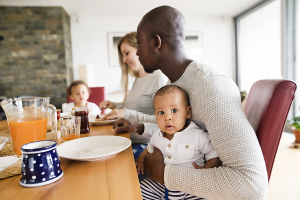 Beautiful young interracial family at home with their cute daughter and little baby son having breakfast together.