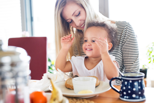 Beautiful young mother at home with his little mixed-race daughter sitting on her lap, having breakfast together.