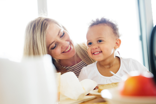 Beautiful young mother at home with his little mixed-race daughter sitting on her lap, having breakfast together.