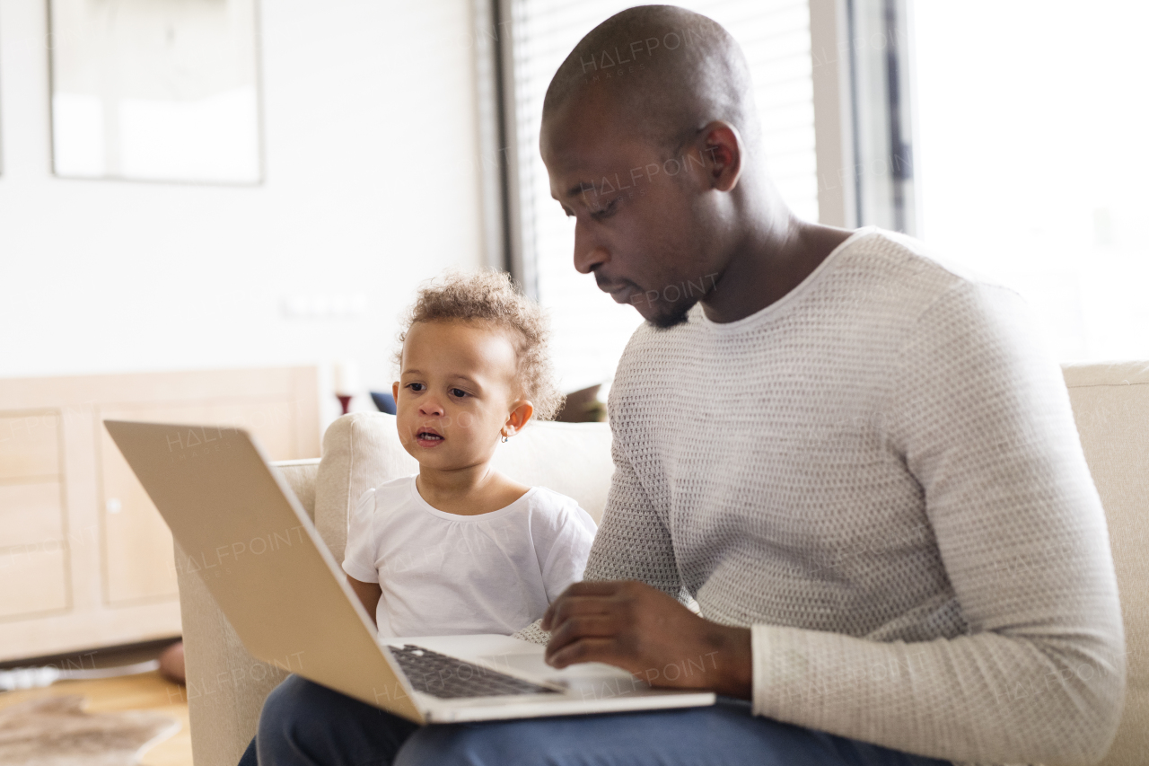 Young afro-american father at home with his cute little daughter sitting on couch watching something on laptop.