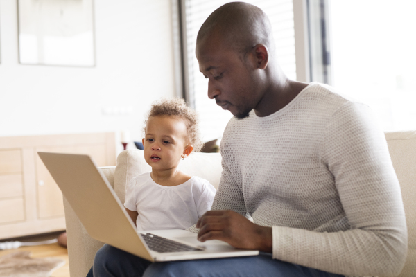 Young afro-american father at home with his cute little daughter sitting on couch watching something on laptop.