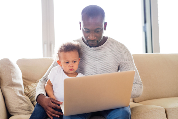 Young afro-american father at home with his cute little daughter sitting on couch watching something on laptop.