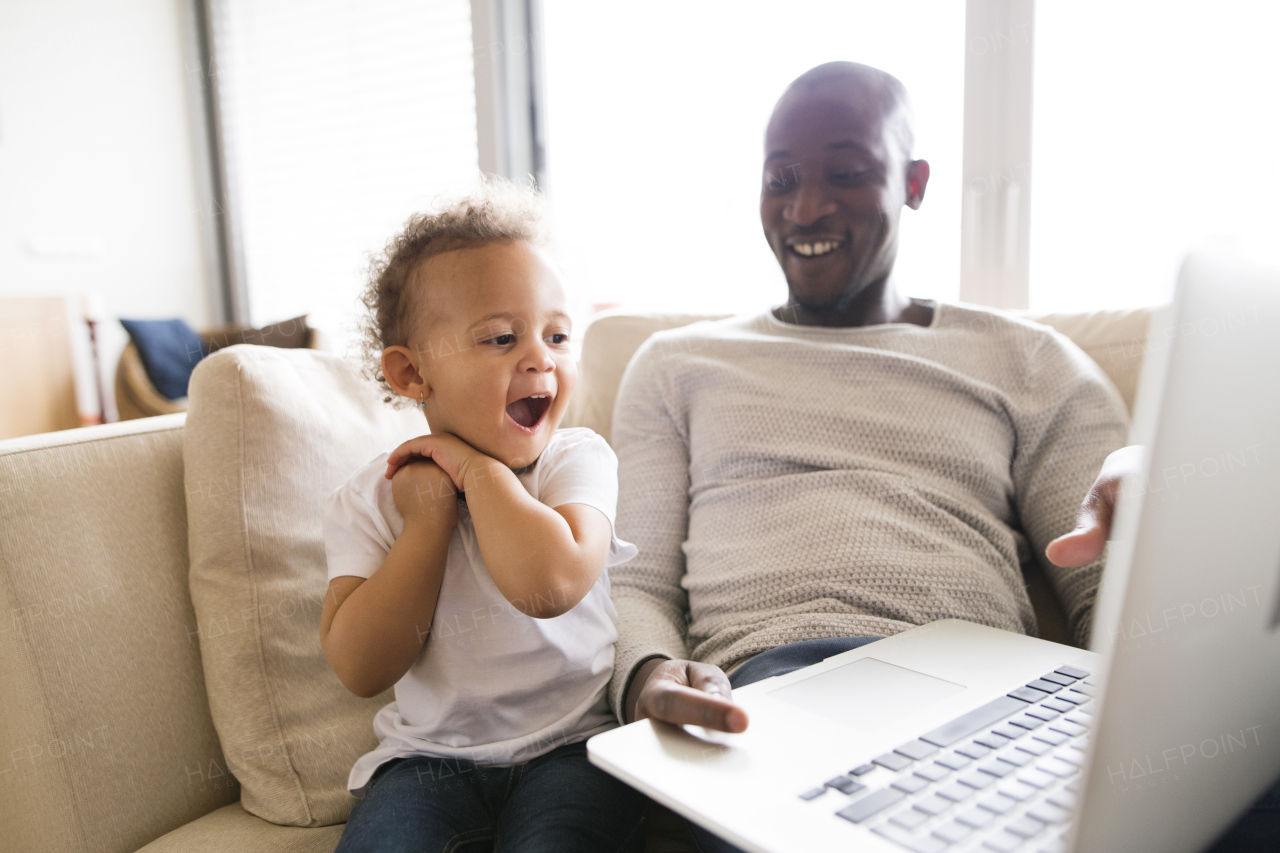 Young afro-american father at home with his cute little daughter sitting on couch watching something on laptop.