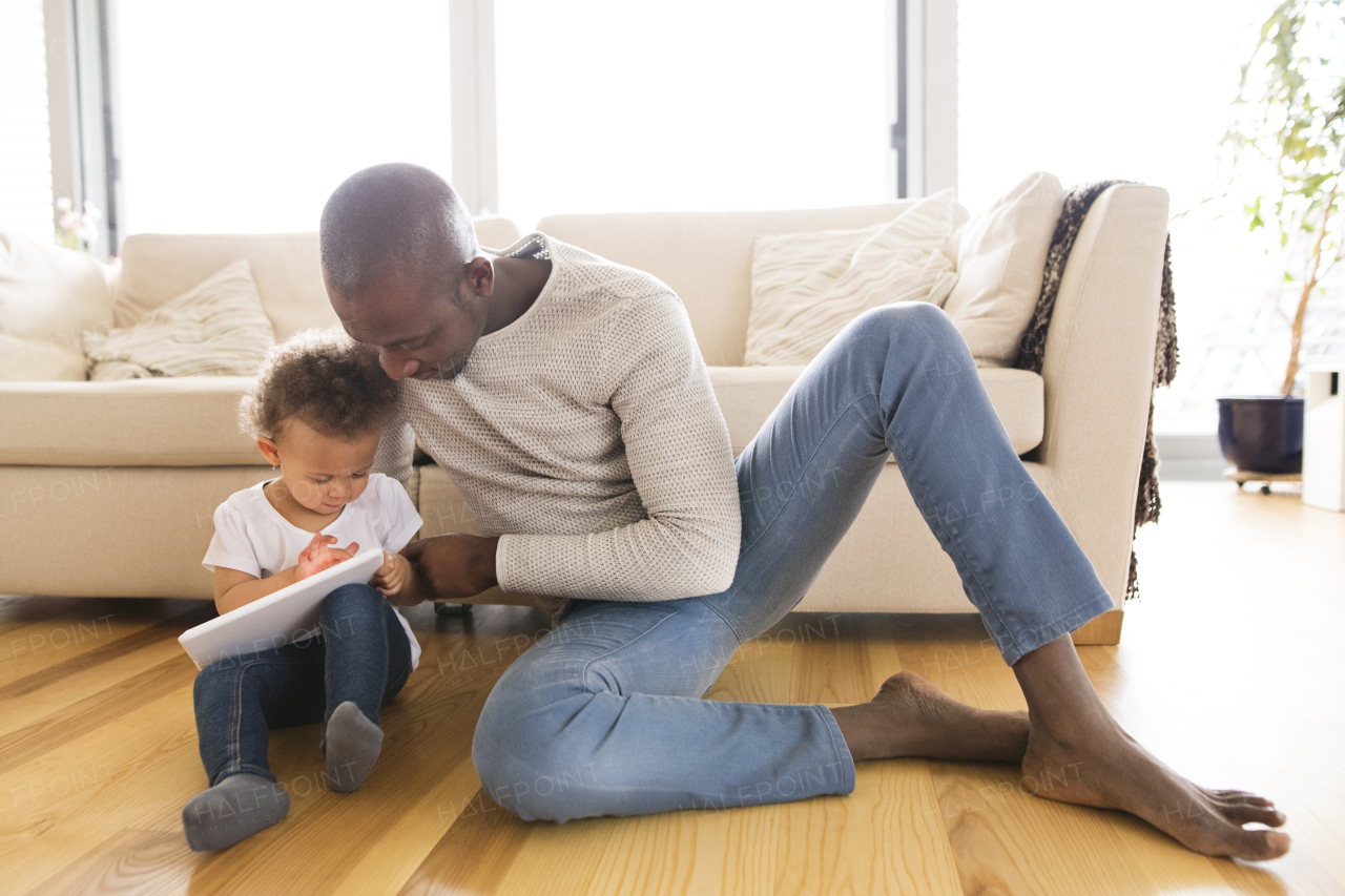 Young afro-american father at home with his cute little daughter sitting on the floor watching something on tablet.